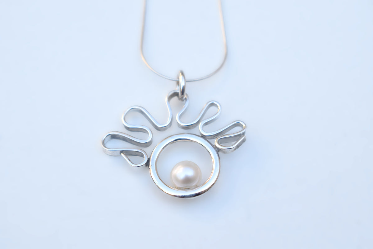 A silver pendant necklace featuring a stylized wavy openwork design surrounding a circular silver frame with a single white pearl, shown on a light blue background.