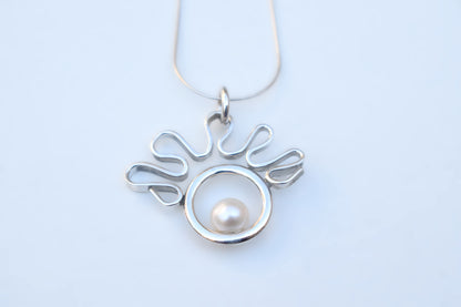 A silver pendant necklace featuring a stylized wavy openwork design surrounding a circular silver frame with a single white pearl, shown on a light blue background.
