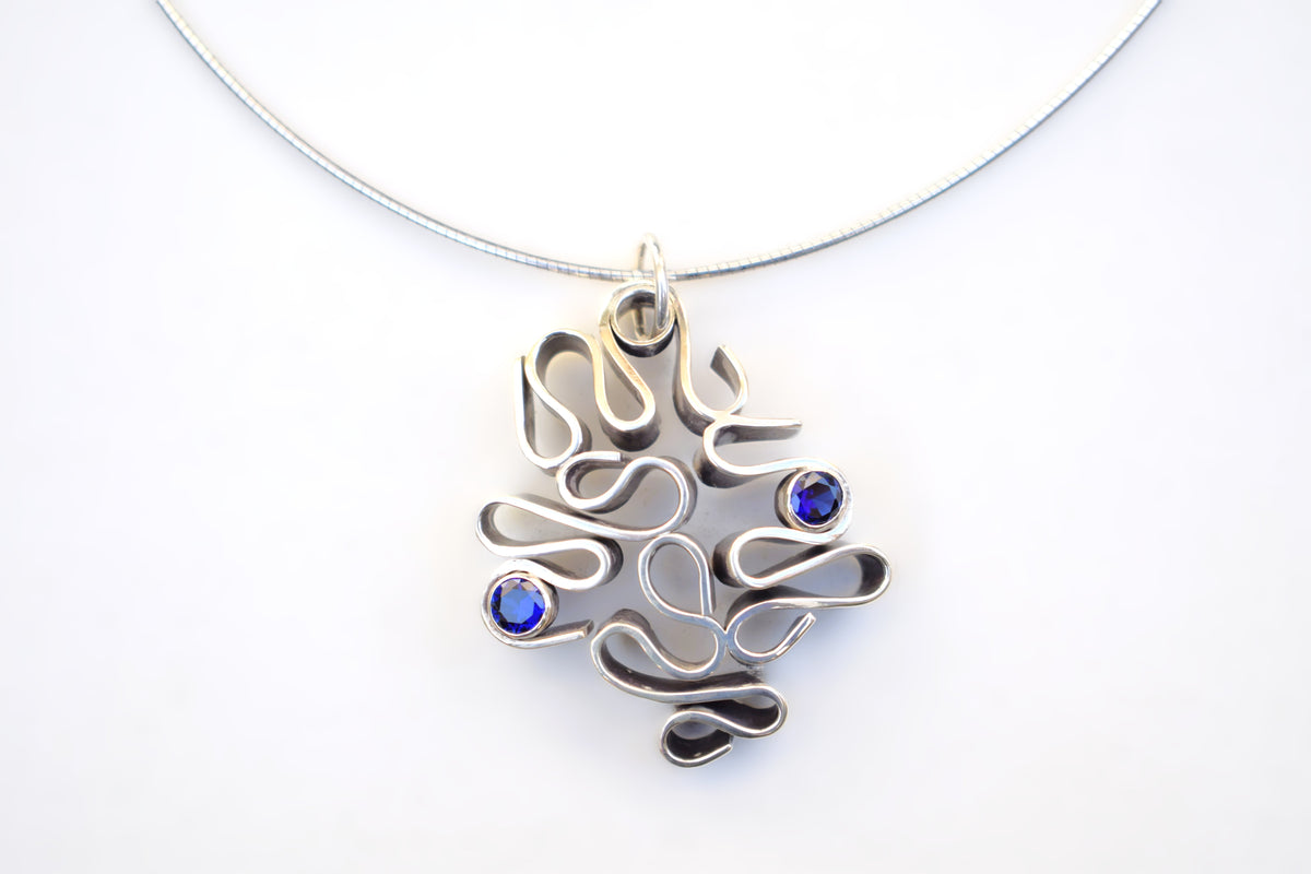 A silver-toned abstract pendant necklace with three small round blue gemstones, shown on a plain white background.