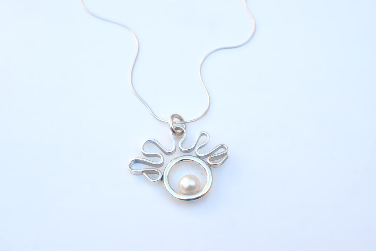 A silver pendant necklace featuring a circular openwork design with a single white pearl in the center, suspended from a delicate silver chain against a light blue background.
