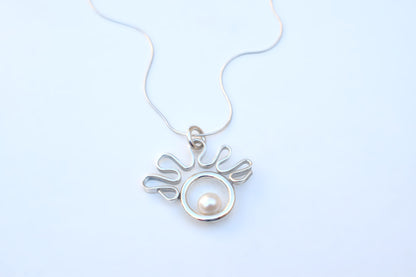 A silver pendant necklace featuring a circular openwork design with a single white pearl in the center, suspended from a delicate silver chain against a light blue background.
