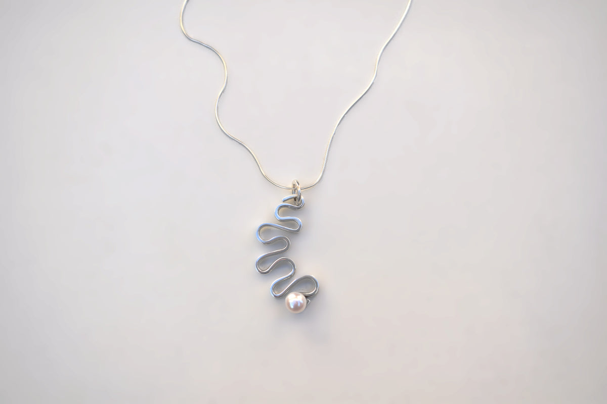 A delicate silver necklace featuring a unique wavy spiral pendant with a small, lustrous white pearl accent, displayed on a clean, light gray background.
