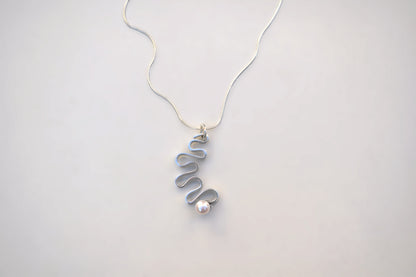 A delicate silver necklace featuring a unique wavy spiral pendant with a small, lustrous white pearl accent, displayed on a clean, light gray background.
