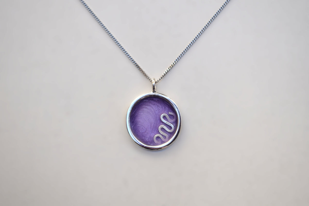 A silver chain necklace with a round bezel pendant containing a purple textured insert with a white snake design, shown on a light gray background.