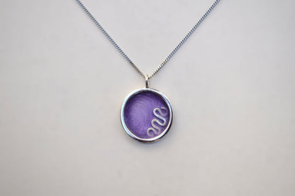 A silver chain necklace with a round bezel pendant containing a purple textured insert with a white snake design, shown on a light gray background.