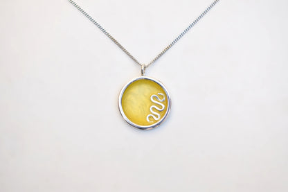 A silver chain necklace with a round pendant featuring a textured yellow center and a white snake design, displayed on a clean white background.