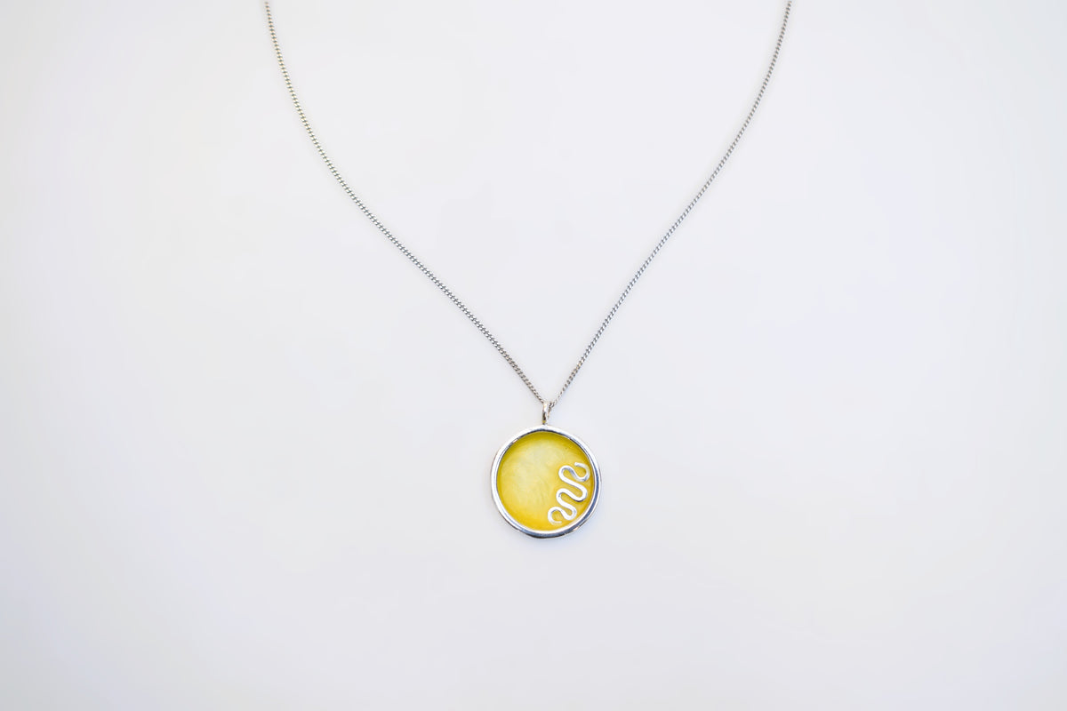 A silver chain necklace with a round pendant featuring a yellow background and a white coiled snake design, displayed against a plain white background.