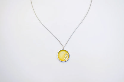 A silver chain necklace with a round pendant featuring a yellow background and a white coiled snake design, displayed against a plain white background.