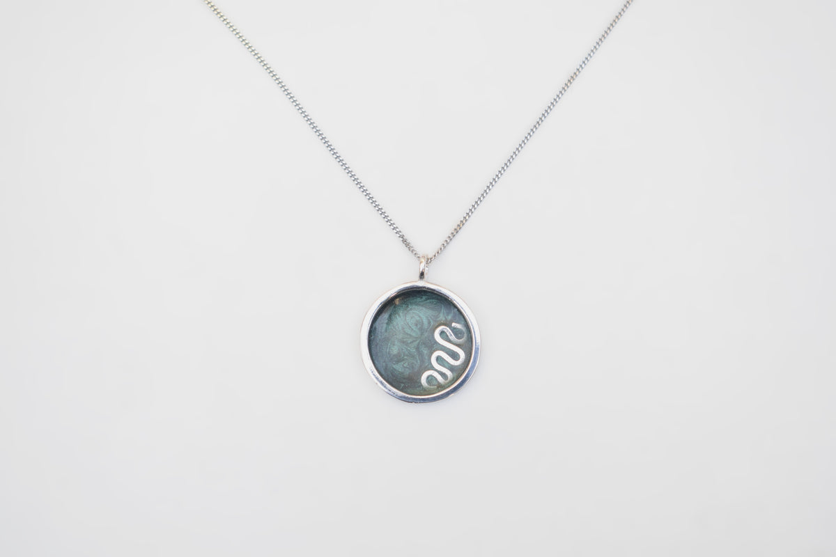 A silver chain necklace with a round bezel pendant containing a dark green-blue stone and a white snake illustration, shown on a plain white background.