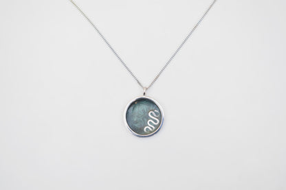A silver chain necklace with a round bezel pendant containing a dark green-blue stone and a white snake illustration, shown on a plain white background.