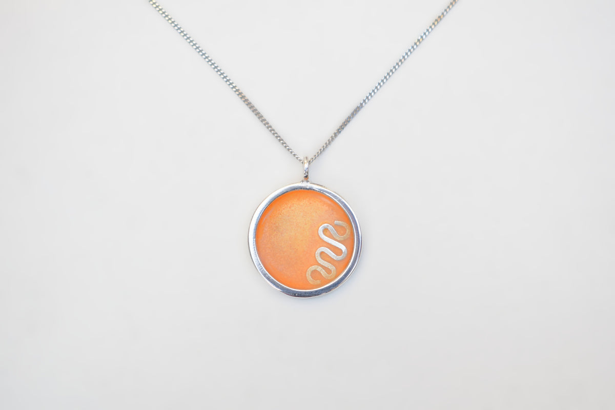 A silver chain necklace with a circular pendant featuring a textured orange background and a white snake design, displayed against a plain white background.