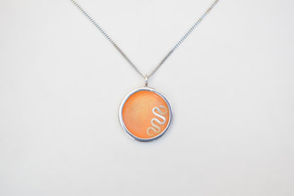 A silver chain necklace with a circular pendant featuring a textured orange background and a white snake design, displayed against a plain white background.