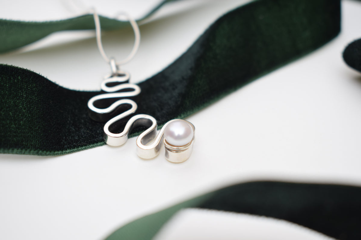 A close-up of a silver wavy pendant necklace with a small pearl bead, draped over a dark green velvet ribbon on a white background.