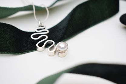 A close-up of a silver wavy pendant necklace with a small pearl bead, draped over a dark green velvet ribbon on a white background.