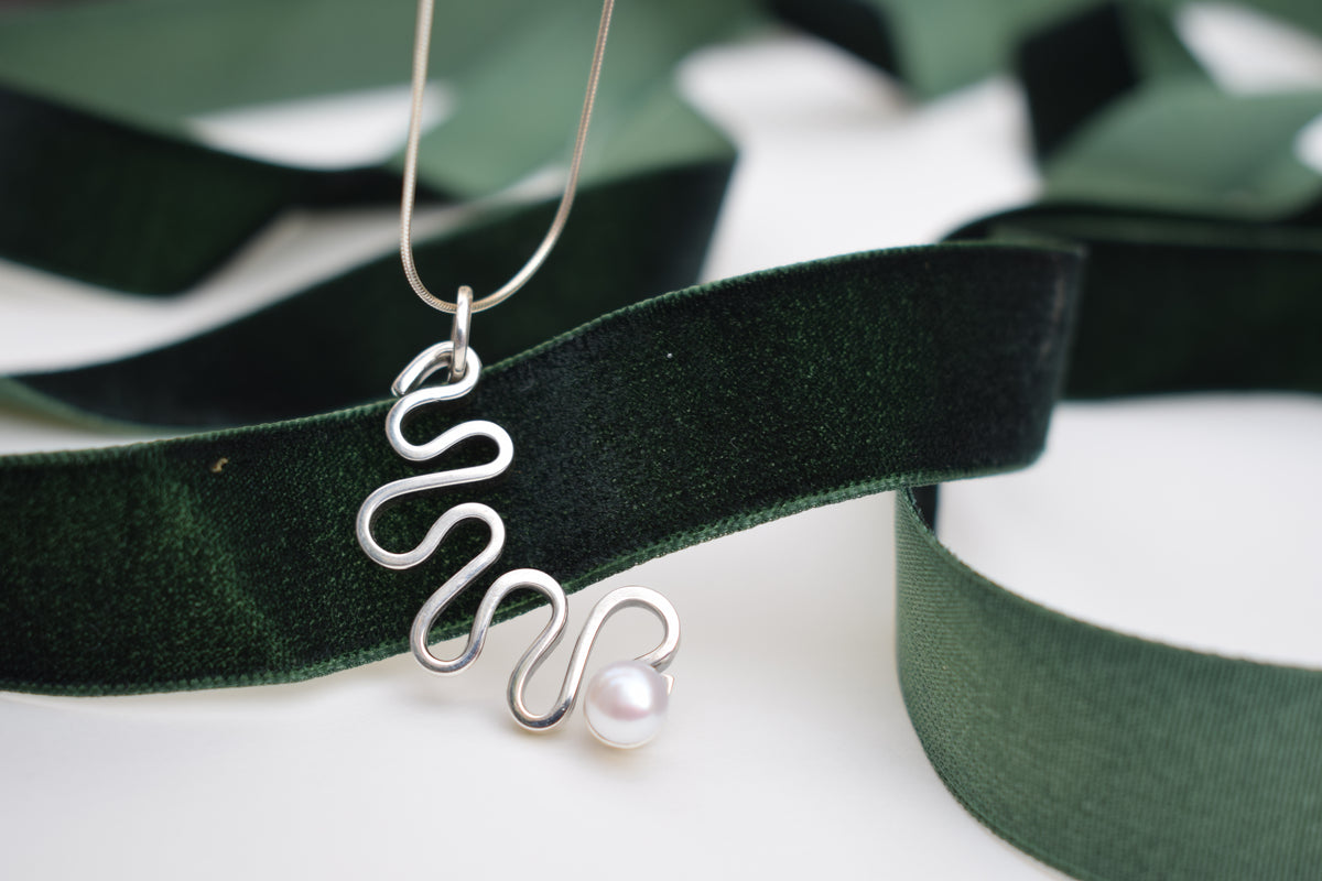 A silver wavy pendant necklace with a single white pearl, draped elegantly over a dark green velvet ribbon on a white background.
