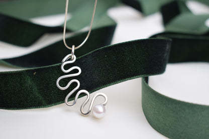 A silver wavy pendant necklace with a single white pearl, draped elegantly over a dark green velvet ribbon on a white background.